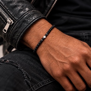 May include: A beaded bracelet featuring small, matte black spherical beads arranged in a continuous line. The bracelet is accented with a small, silver-toned metal cross charm positioned centrally, flanked by two tiny silver-toned metal spacer beads.