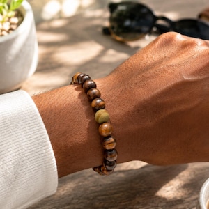 Könnte beinhalten: Ein Perlenarmband mit dunkelbraunen Holzperlen und einer einzelnen, zentralen hell olivgrünen Perle. Die Perlen sind kugelförmig und auf ein elastisches Band gefädelt, wodurch ein kreisförmiges Band entsteht, das um das Handgelenk passt.