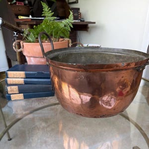 May include: A large, antique copper pot with two small handles. The pot has a wide, rounded shape and a shiny, reflective surface. The pot is sitting on a glass table. A stack of blue books and a terracotta pot with a fern are in the background.