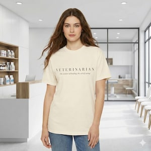 May include: Cream-colored t-shirt with the word "VETERINARIAN" printed in black, along with the text "The science of healing, the art of caring." The shirt is worn with blue jeans. The background is a veterinary clinic.