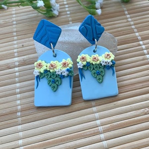 Closeup image of earrings.  Textured leaf design in the top part of the post earrings can be seen as well as intricate details of the yellow flowers with orange centers, tiny white blossoms, green cascading leaves and blue berry like accents.