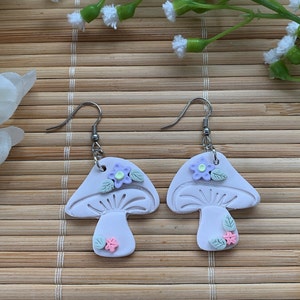 Closeup of earrings shown against a neutral beige background. Details such as tiny green leaves, purple flower with yellow center and itty bitty pink flower can be seen.