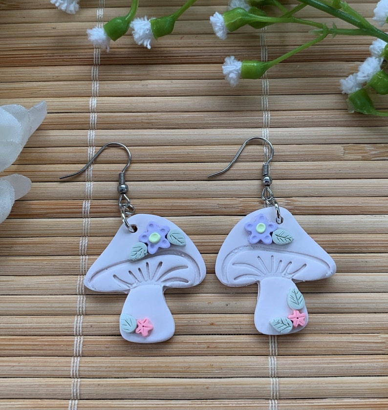 Closeup of earrings shown against a neutral beige background. Details such as tiny green leaves, purple flower with yellow center and itty bitty pink flower can be seen.