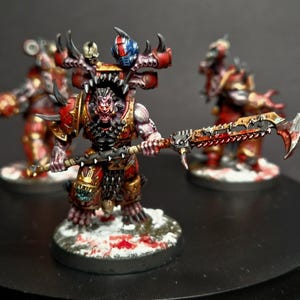May include: A red and black painted miniature figure of a Chaos Space Marine wielding a chain sword. The figure is standing on a snowy base with blood splatters.