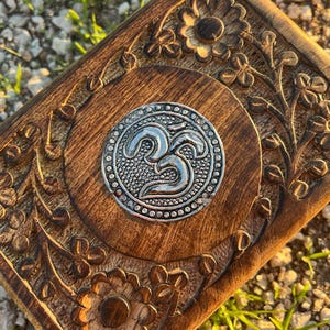 Puede incluir: Una caja de madera con intrincados tallados florales y un símbolo Om plateado en el centro. La caja es de color marrón oscuro y tiene un acabado pulido.