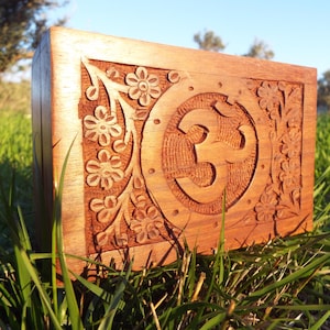 Puede incluir: Una caja de madera con intrincados tallados de flores y un símbolo Om. La caja es de color marrón oscuro y está sentada sobre un lecho de hierba verde.