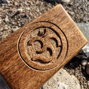Puede incluir: Una caja de madera con un símbolo Om tallado en un círculo en la parte superior. La caja está hecha de madera marrón oscura y tiene un acabado natural.