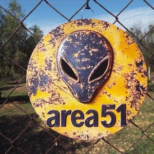 Può includere: Un cartello rotondo e usurato con sfondo giallo e marrone, raffigurante una testa aliena scura e la scritta "area 51" in nero. Il cartello è appeso a una recinzione a rete metallica, con cielo blu e alberi sullo sfondo.