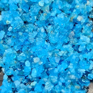 May include: A close-up of a collection of small, irregularly shaped, translucent blue crystals. The crystals vary in size and have a slightly rough texture, with some lighter, almost white, inclusions.