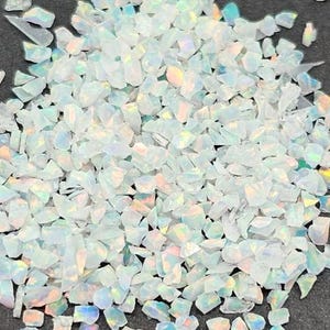 May include: A pile of small, irregularly shaped, white opal chips. The chips have a translucent quality, with flashes of iridescent color visible throughout. The chips are scattered across a dark surface.