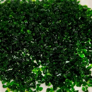 May include: Close-up of a pile of small, irregularly shaped, dark green glass pieces. The pieces are scattered across a white surface, creating a textured, sparkling appearance. The glass appears to be crushed or broken.