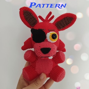 Könnte beinhalten: Ein handgefertigtes rotes Häkelfuchs-Spielzeug mit einer schwarzen Augenklappe und einem gelb-weißen Auge. Der Fuchs hat braune Ohren und hält einen kleinen weißen Zahn. Der Text "ENGLISH PDF PATTERN" wird oben im Bild angezeigt.