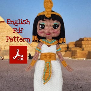 May include: A handmade crochet doll depicting an Egyptian queen, wearing a white dress with gold and turquoise accents. The doll has black hair, a gold headdress, and is standing in front of a pyramid. The image includes the text "English Pdf Pattern".