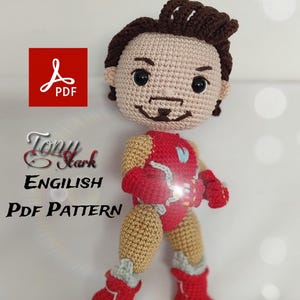 May include: Crocheted Tony Stark doll in a standing pose. The doll features a tan face with brown hair, a red and gold suit, and red boots. The text "Tony Stark ENGLISH PDF PATTERN" is visible.