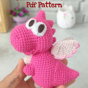 Puede incluir: Juguete de amigurumi de dragón de crochet rosa con ojos blancos y pupilas marrones. El dragón tiene alas rosas y una cola. El juguete se sostiene en una mano.