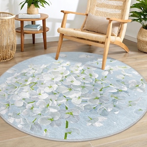 Puede incluir: Una alfombra redonda de color azul claro con un diseño floral blanco. La alfombra presenta un gran grupo de flores blancas con tallos y hojas verdes. La alfombra es perfecta para agregar un toque de elegancia y naturaleza a cualquier habitación.