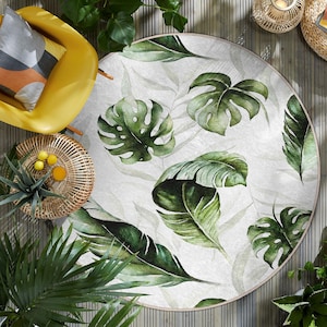 Könnte beinhalten: Ein runder, weißer Teppich mit einem grünen tropischen Blattmotiv. Der Teppich hat einen dezenten Aquarelleffekt und zeigt große, detaillierte Blätter.