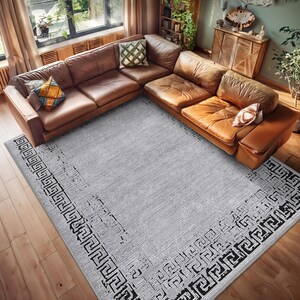 Könnte beinhalten: Ein grauer Teppich mit einem schwarzen und weißen geometrischen Rand. Der Teppich befindet sich in einem Wohnzimmer mit einem braunen Ledersofa.