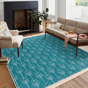Op de afbeelding: Een tealblauw vloerkleed met een wit pijlpatroon. Het kleed heeft een crèmekleurige rand en ligt op een houten vloer in een woonkamer.