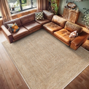 May include: A natural fibre rug in a light beige colour with a subtle woven pattern. The rug is placed on a hardwood floor in a living room setting with a brown leather sectional sofa.