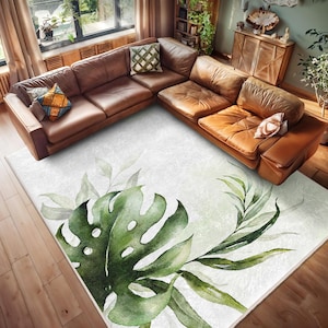 May include: A white area rug with a watercolour design of green leaves and a large monstera leaf. The rug is placed in a living room with a brown leather sectional sofa.