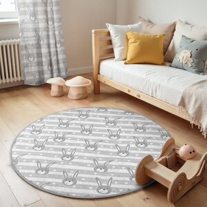 May include: A round gray and white play mat with a pattern of gray bunny faces and white stars on gray stripes. The mat is on a wooden floor in a room with a wooden daybed and a gray and white patterned curtain.