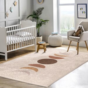 Könnte beinhalten: Ein beiger Teppich mit einem Muster aus braunen und orangen Halbmonden. Der Teppich befindet sich in einem Kinderzimmer mit einem weißen Babybett, einer Topfpflanze und einem weißen Hocker.