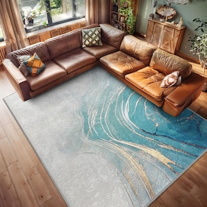 May include: A blue and white abstract area rug with gold accents. The rug is laid out on a wooden floor in a living room with a brown leather sectional sofa.