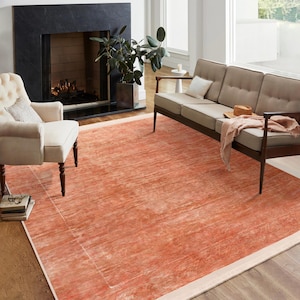 Op de afbeelding: Een groot, rechthoekig, oranje-rood tapijt met een subtiel, vervaagd patroon en een witte rand. Het tapijt bevindt zich in een woonkamer met een open haard en een bank.