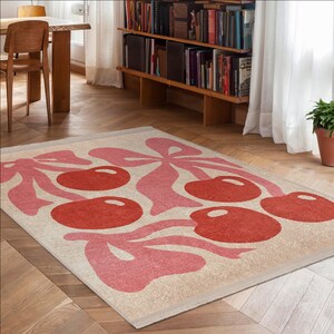 May include: A cream-coloured rug with a pink and red cherry design. The rug has a fringe border and is laid out on a hardwood floor.