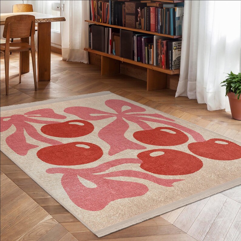 May include: A cream-coloured rug with a pink and red cherry design. The rug has a fringe border and is laid out on a hardwood floor.