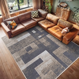 以下が含まれることがあります： グレーとベージュの幾何学模様のエリアラグ。ラグは、グレーとベージュのさまざまな色合いの正方形と長方形で構成されたモダンなデザインです。ラグは、リビングルームの硬材の床に敷かれています。