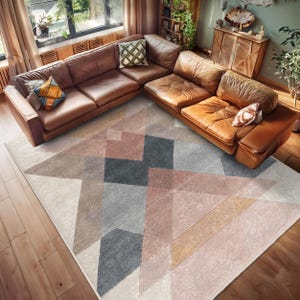 May include: A geometric patterned rug with shades of pink, gray, and beige. The rug is laid out on a hardwood floor in a living room setting with a brown leather sectional sofa.
