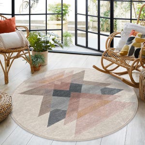Puede incluir: Una alfombra redonda de color crema con un patrón geométrico de triángulos rosas, naranjas y grises. La alfombra está en un salón con una mecedora de mimbre y una planta en maceta.