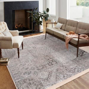 May include: A light grey area rug with a patchwork design featuring various patterns in shades of grey and brown. The rug has a cream-coloured border and is displayed in a living room setting with a sofa and armchair.