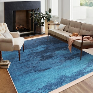 May include: A blue and teal abstract patterned area rug with a cream border. The rug is in a living room setting with a couch and a chair.
