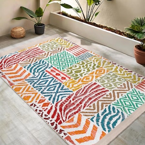 Op de afbeelding: Veelkleurig vloerkleed met een patchwork-ontwerp met verschillende geometrische patronen, waaronder strepen, zigzagpatronen en ruiten. Het kleed heeft een witte achtergrond en een beige rand.