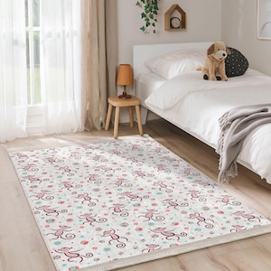 Può includere: Un tappeto bianco con un motivo di scimmia rosa e blu. Il tappeto è su un pavimento bianco in una camera da letto con un letto bianco e un tavolino di legno.