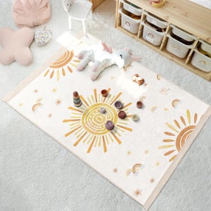 Puede incluir: Una alfombra de juego blanca y beige con un diseño de sol y arcoíris. La alfombra presenta múltiples soles con acentos amarillos y naranjas y arcoíris en tonos azules y rosas. La alfombra es perfecta para la sala de juegos o la habitación de un niño.