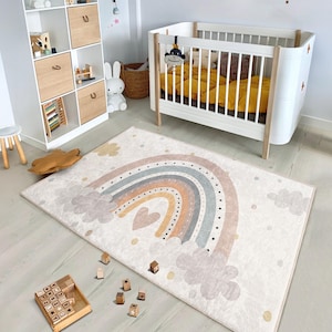 Puede incluir: Una alfombra de arcoíris blanca y beige con nubes y puntos dorados. La alfombra presenta un gran arcoíris con un corazón en el centro. La alfombra está en una guardería con una cuna blanca y estantes de madera.