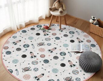 Puede incluir: Una alfombra de juego redonda de color crema con un borde rosa presenta un patrón repetitivo de planetas, estrellas y cohetes en tonos de azul, verde y rosa.