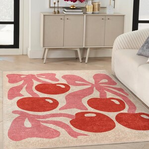 May include: A pink and red area rug with a cherry design. The rug has a cream background with pink cherry stems and leaves and red cherries.