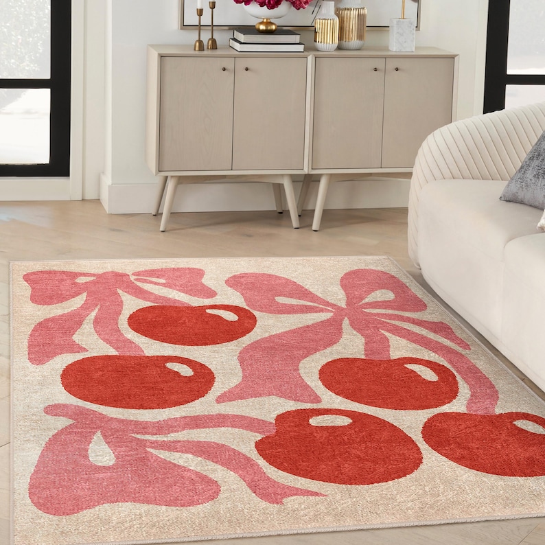 May include: A pink and red area rug with a cherry design. The rug has a cream background with pink cherry stems and leaves and red cherries.