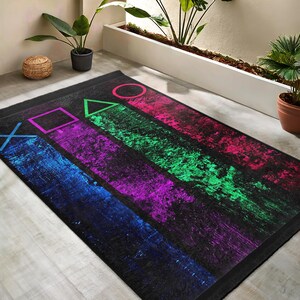 May include: A black rug with a colorful design featuring the PlayStation controller symbols. The symbols are in green, blue, purple, pink, and red. The rug has a distressed, textured look.