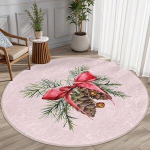 Könnte beinhalten: Ein runder rosa Teppich mit einem Aquarell-Design aus einem Kiefernzweig mit einer roten Schleife und Tannenzapfen.