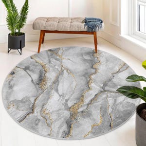 Puede incluir: Una alfombra redonda gris y blanca con un patrón de mármol y detalles dorados. La alfombra tiene un brillo sutil.