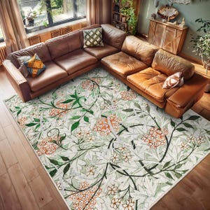 May include: A white area rug with a floral pattern featuring green leaves and orange flowers. The rug is laid out on a wooden floor in a living room setting with a brown leather sectional sofa.