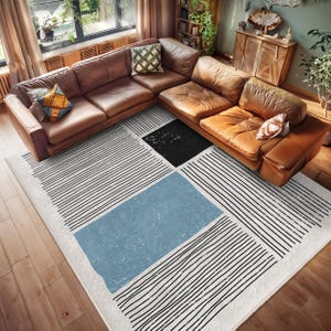 May include: A white and black striped rug with a blue rectangle and a black square in the center. The rug is on a wooden floor in front of a brown leather sectional sofa.