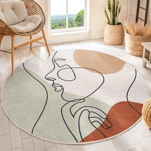 Può includere: Un tappeto rotondo con un design astratto che presenta un disegno a linea di un viso con una mano sul mento. Il tappeto ha uno sfondo beige con accenti marroni, arancioni e verdi.