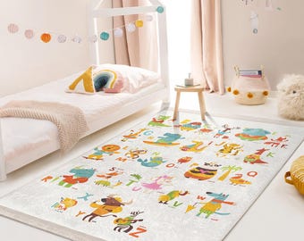 Op de afbeelding: Een wit en grijs alfabetkleed met kleurrijke dierillustraties. Elk dier houdt een letter van het alfabet vast. Het kleed ligt op een witte vloer in een kinderkamer.